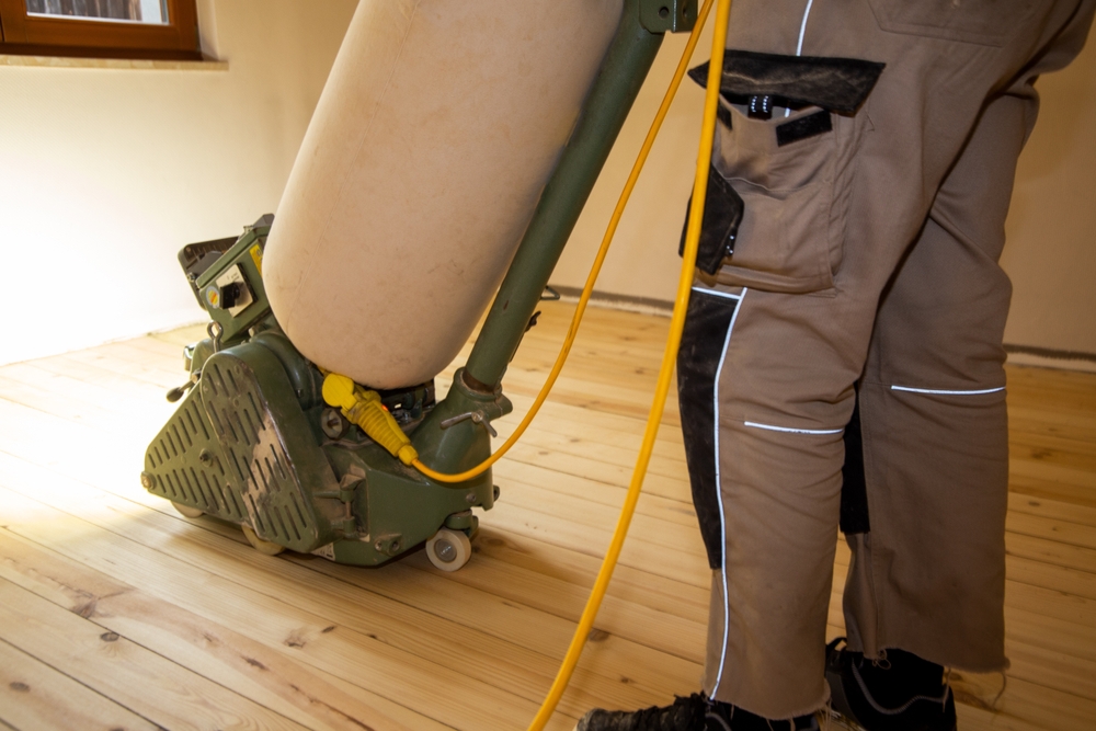 A close-up of a professional using a sanding machine on a solid pine wooden floor in West Kensington, with a dust collection bag attached, during a wood floor sanding project.