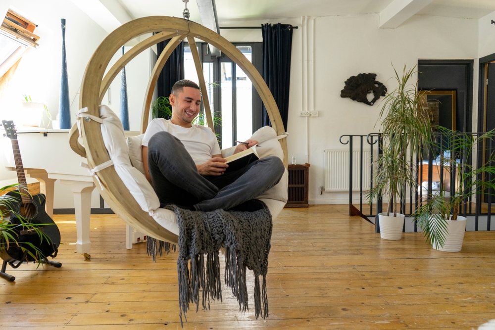 A person relaxing in a hanging egg chair in a West Kensington room, reading a book on a beautifully sanded solid oak wooden floor, surrounded by plants, a guitar, and a piano.