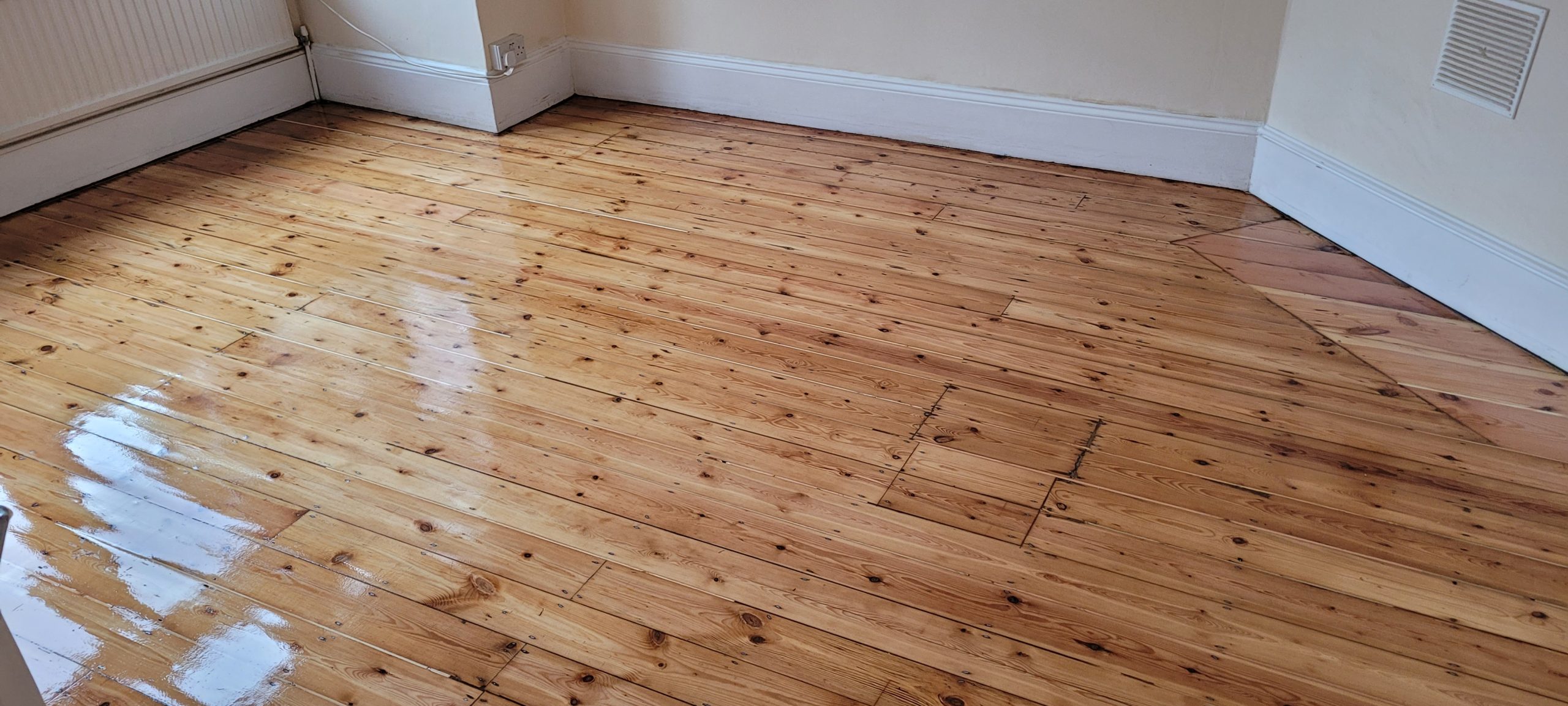 A beautifully polished solid oak wooden floor in a West Kensington room, showcasing the glossy finish achieved through professional wood floor sanding, with white baseboards and a radiator in the background.