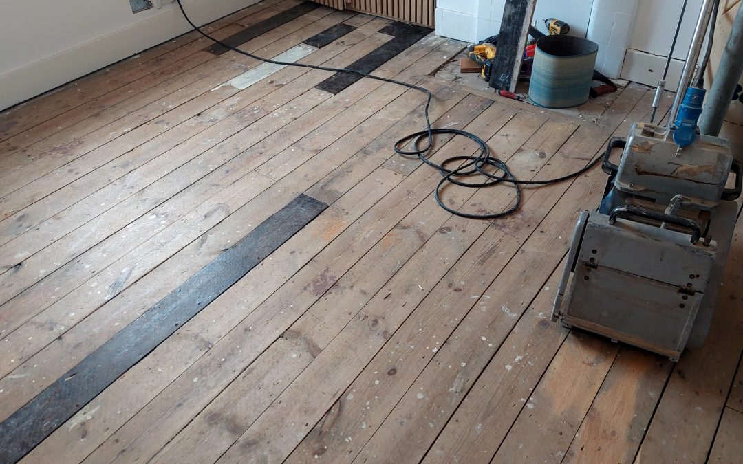 A room in West Kensington during wood floor sanding, featuring a solid pine wooden floor with a sanding machine, tools, and dust collection equipment, showing the initial stage of restoration.