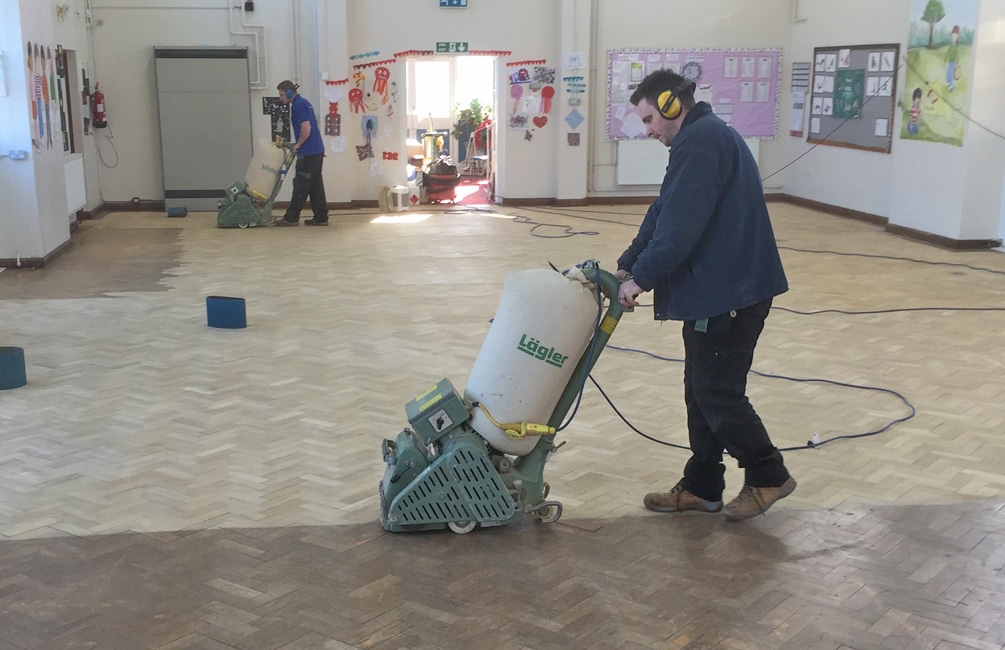 Parquet Floor Sanding in West Kensington – contractors sanding an oak herringbone parquet school hall floor with professional dust-controlled machines.